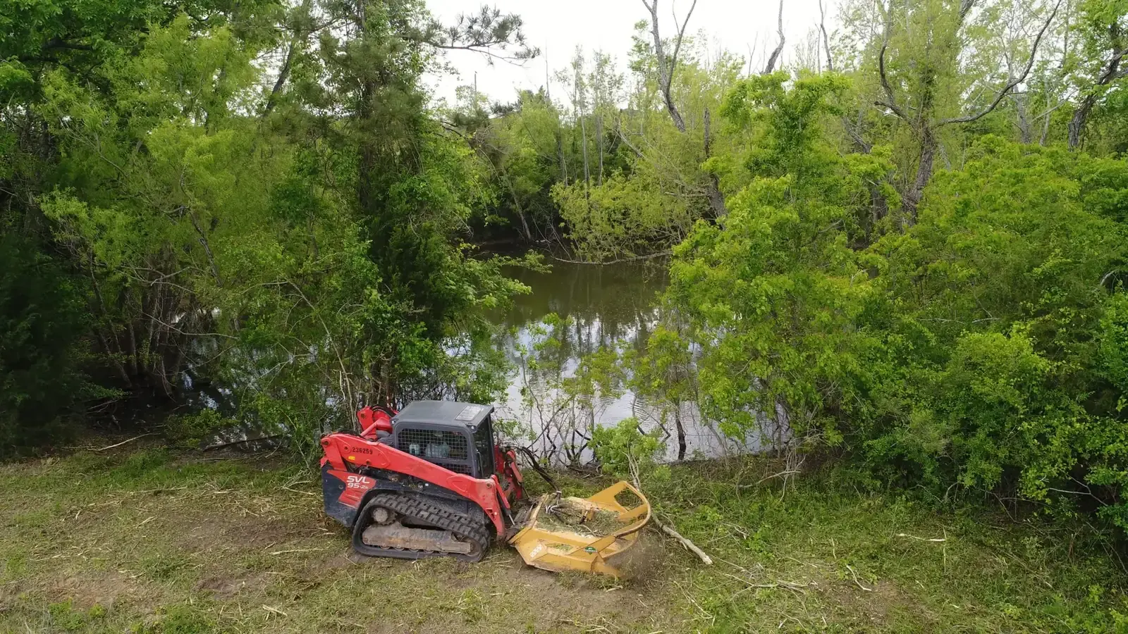 Track loader with a mulching head clearing brush along a pond edge