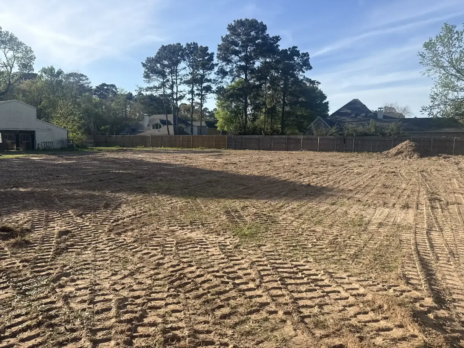 Graded dirt lot with equipment tracks beside a tree-lined fence