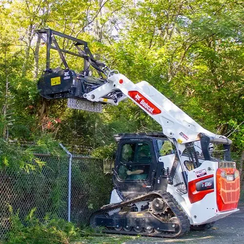 Bobcat mulcher with boom raised trimming overgrowth along a chain-link fence