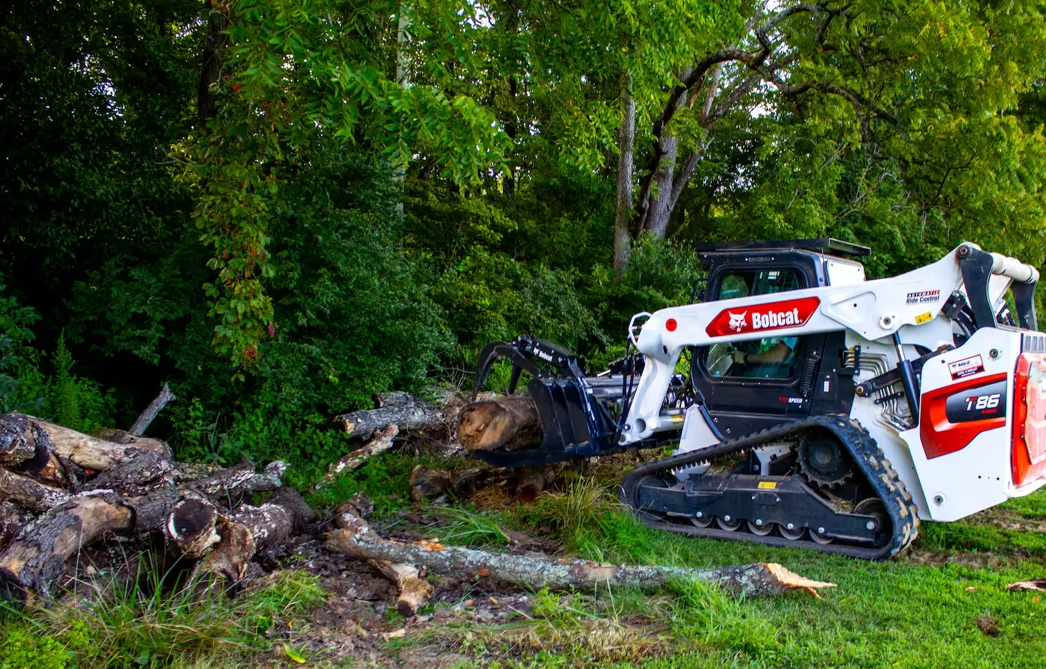 Bobcat T86 with grapple moving cut logs beside a wooded edge