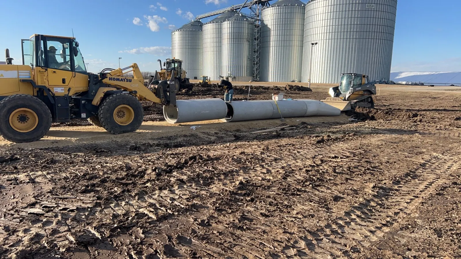 Large drainage pipe being set beside grain bins with a wheel loader and dozer